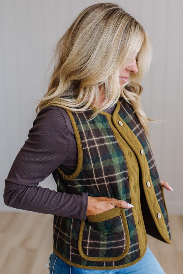 A woman wearing a green and black plaid quilted vest with olive green trim over a dark plum long-sleeve shirt and blue jeans, shown in a side profile against a neutral indoor background.