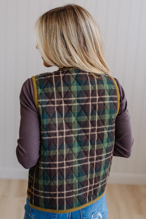 A woman viewed from behind wearing a quilted plaid vest over a dark purple long-sleeve shirt and blue jeans, standing in a brightly lit indoor room with a white paneled wall and light wood floor.