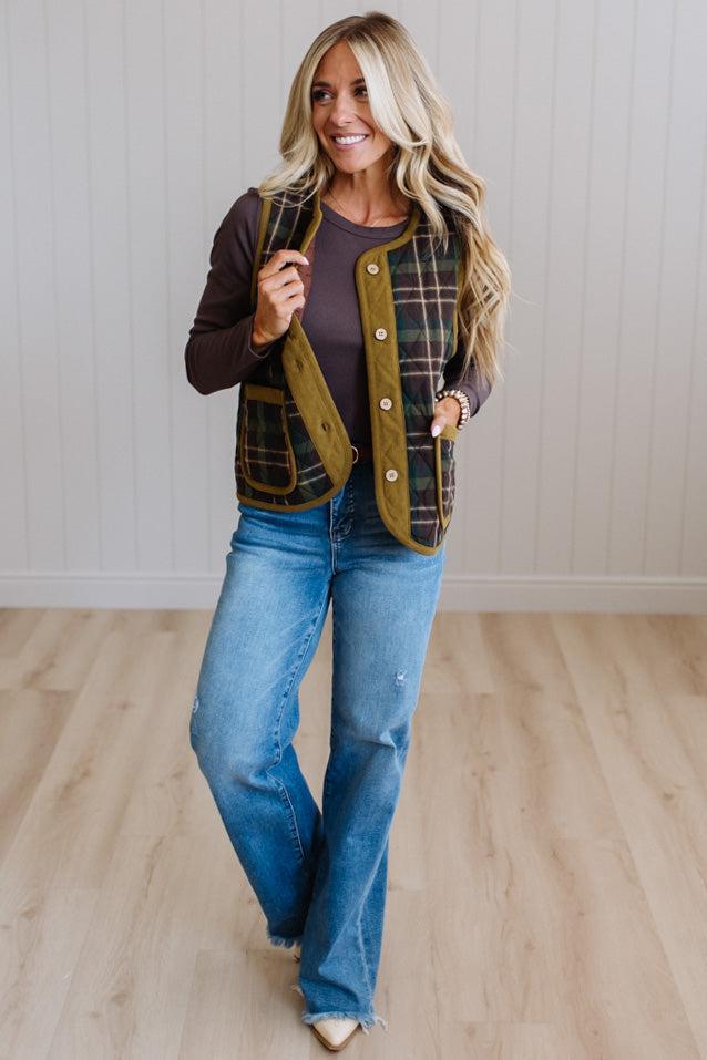 A woman modeling a green and black plaid button-up vest over a dark brown long-sleeve shirt and blue jeans, standing in a room with light wood floors and white paneled walls.