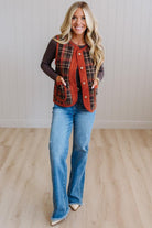 A woman modeling a rust red and black plaid button-up vest over a dark brown long-sleeve shirt and blue jeans, standing in a room with light wood floors and white paneled walls.