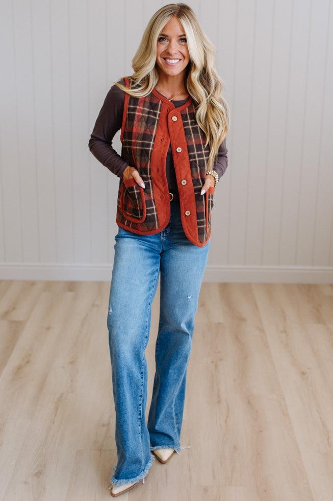 A woman modeling a rust red and black plaid button-up vest over a dark brown long-sleeve shirt and blue jeans, standing in a room with light wood floors and white paneled walls.