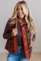 A smiling blonde woman wearing a brown and red plaid button-front vest over a dark brown long-sleeve top and blue jeans, standing against a light paneled wall.