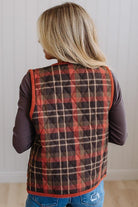 A woman viewed from behind wearing a quilted sleeveless vest with a red, brown, and beige plaid pattern over a dark long-sleeve shirt and blue jeans.