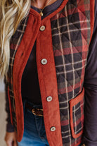 A close-up of a woman wearing a mixed-media vest with a rust orange corduroy front panel and trim, and a red, black, and beige plaid quilted panel on the back. She is wearing blue jeans and a black top underneath.