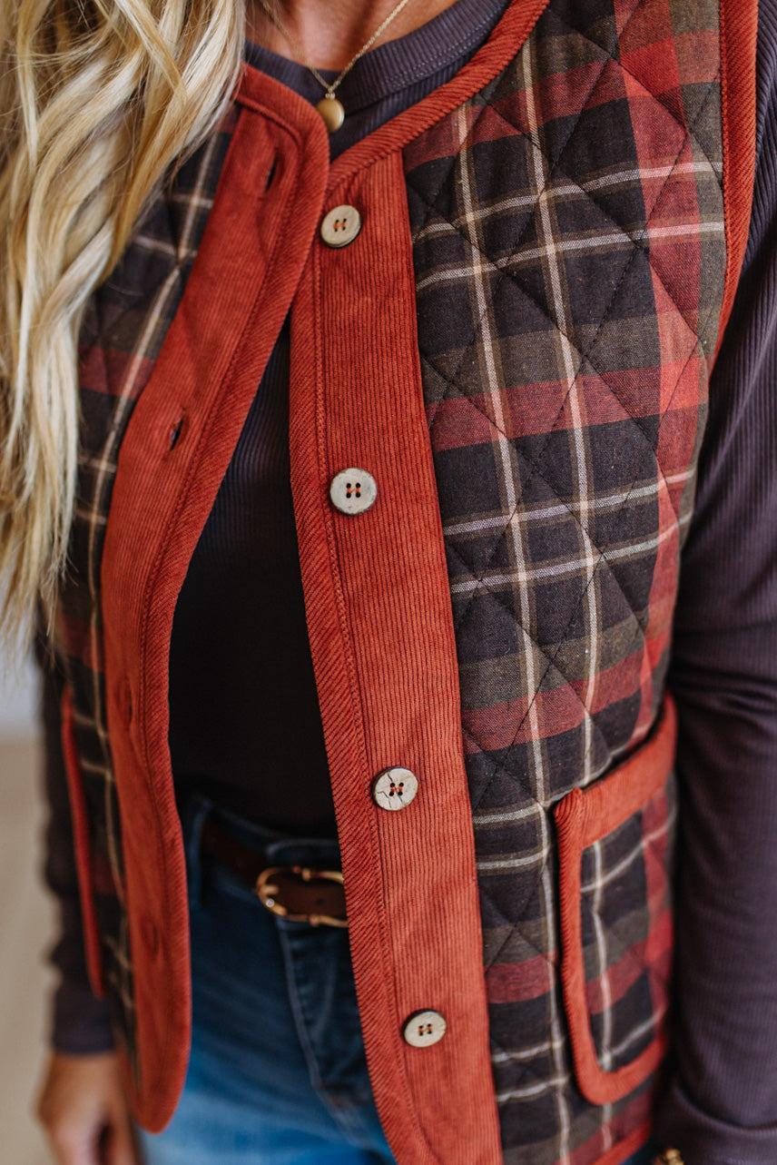 A close-up of a woman wearing a mixed-media vest with a rust orange corduroy front panel and trim, and a red, black, and beige plaid quilted panel on the back. She is wearing blue jeans and a black top underneath.