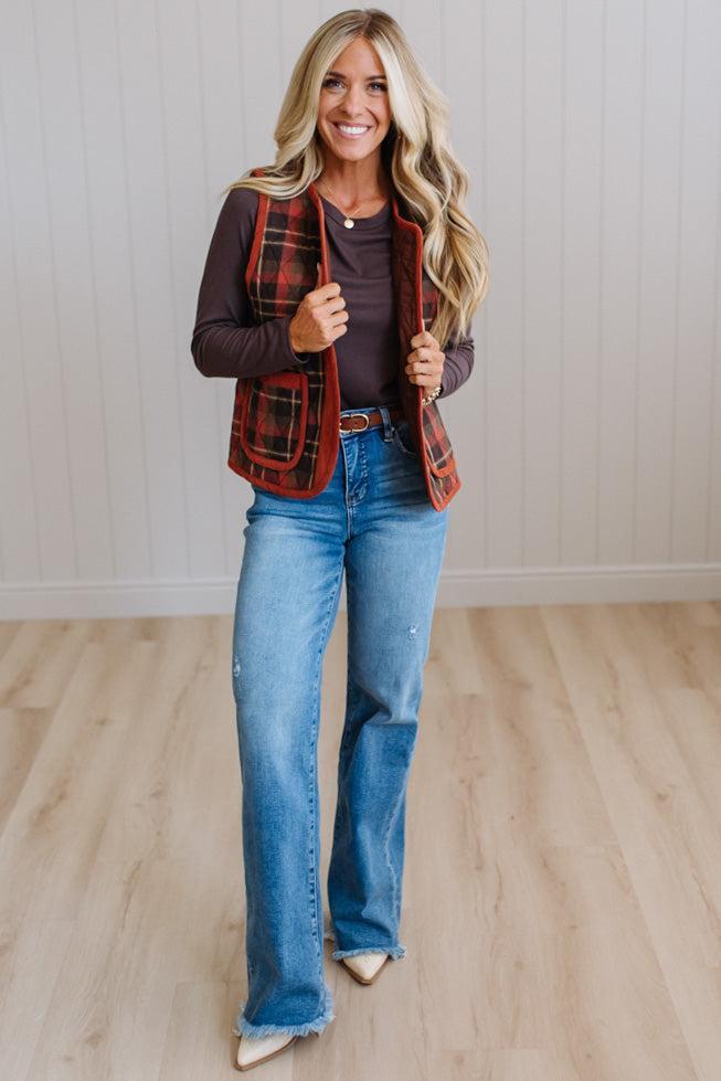 A woman modeling a red and black plaid vest over a dark brown long-sleeve top, paired with blue jeans and beige pointed-toe heels, standing in a room with light wood floors and white paneled walls.
