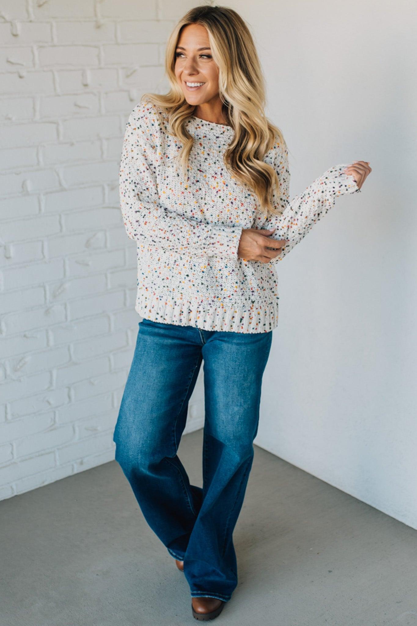 Woman wearing a patterned sweater and blue jeans against a white brick wall.