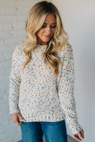 Woman wearing a long-sleeve, patterned top against a white brick wall.