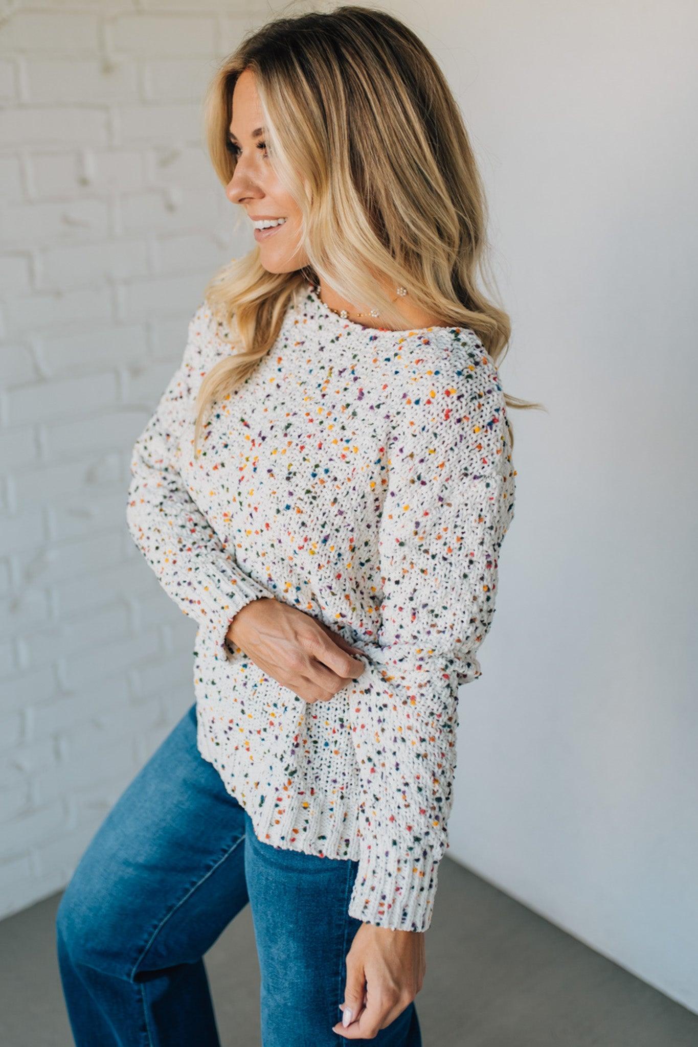 Woman wearing a patterned long-sleeve top and blue jeans against a white brick wall.