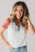 BLONDE MODEL WEARING A COLOR CONTRASTED, HALF BUTTON, SHORT SLEEVE, COLOR BLOCKED BASEBALL TOP IN FADE RED WHITE AND BLUE.