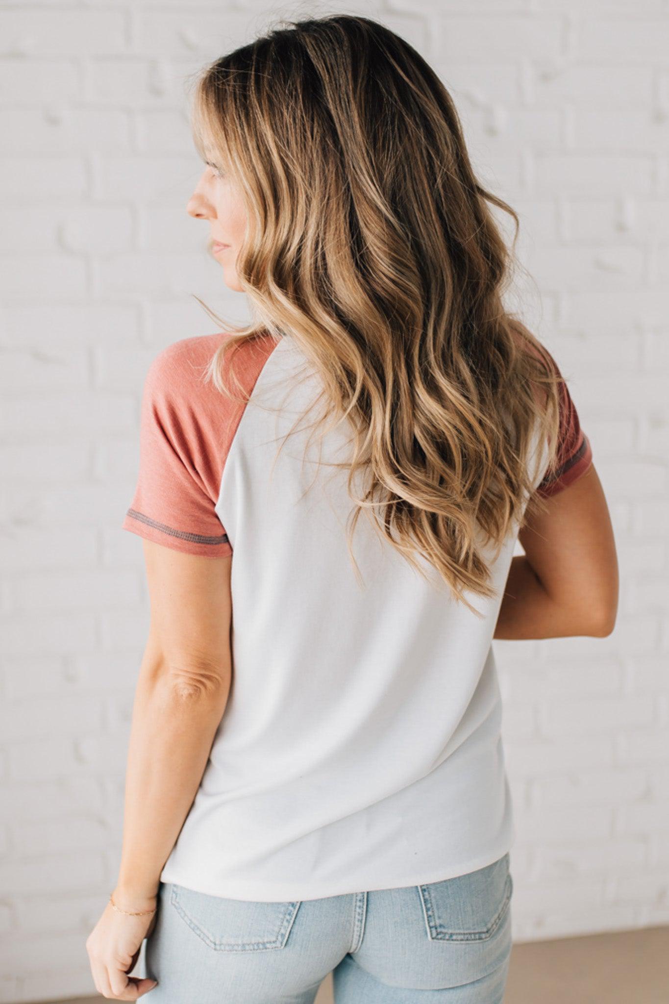 BLONDE MODEL WEARING A COLOR CONTRASTED, HALF BUTTON, SHORT SLEEVE, COLOR BLOCKED BASEBALL TOP IN FADE RED WHITE AND BLUE.