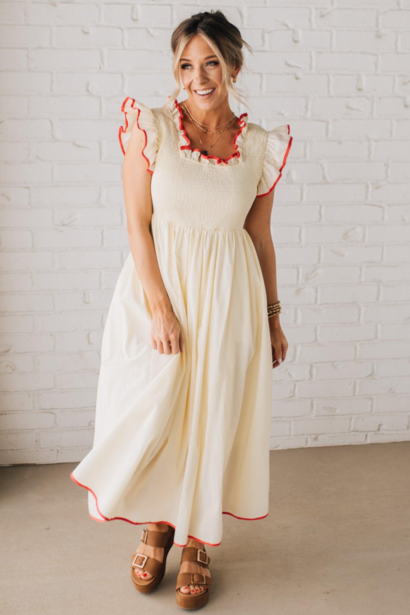 BLONDE MODEL WEARING A CREAM DRESS WITH RED CONTRASTED BINDING DETAIL AND SMOCKED BODICE MIDI DRESS WITH RUFFLE ACCENTS AND TIE BACK DETAIL.