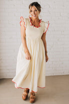 BLONDE MODEL WEARING A CREAM DRESS WITH RED CONTRASTED BINDING DETAIL AND SMOCKED BODICE MIDI DRESS WITH RUFFLE ACCENTS AND TIE BACK DETAIL.