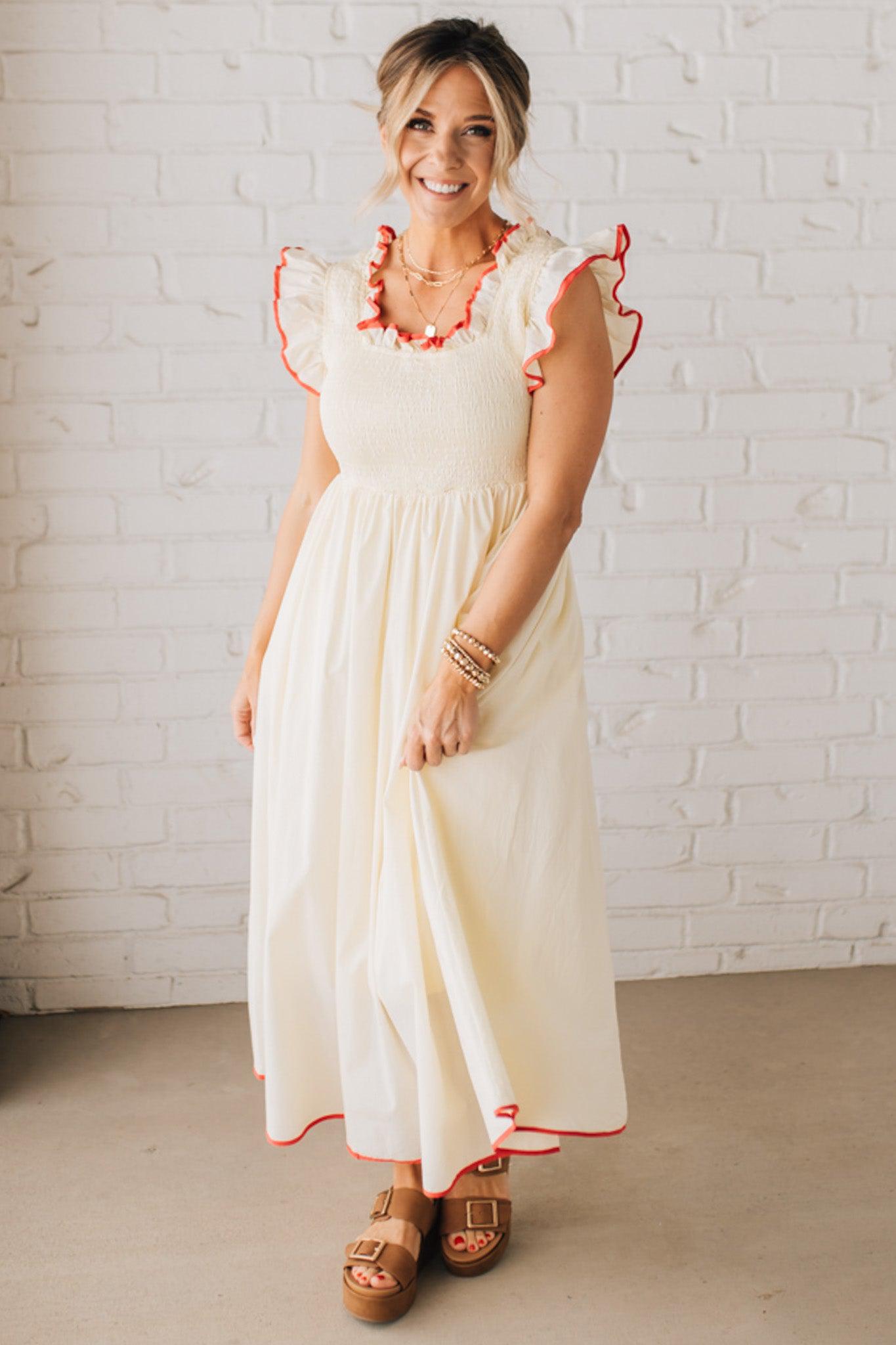 BLONDE MODEL WEARING A CREAM DRESS WITH RED CONTRASTED BINDING DETAIL AND SMOCKED BODICE MIDI DRESS WITH RUFFLE ACCENTS AND TIE BACK DETAIL.