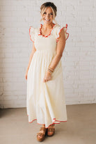 BLONDE MODEL WEARING A CREAM DRESS WITH RED CONTRASTED BINDING DETAIL AND SMOCKED BODICE MIDI DRESS WITH RUFFLE ACCENTS AND TIE BACK DETAIL.