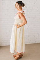 BLONDE MODEL WEARING A CREAM DRESS WITH RED CONTRASTED BINDING DETAIL AND SMOCKED BODICE MIDI DRESS WITH RUFFLE ACCENTS AND TIE BACK DETAIL.