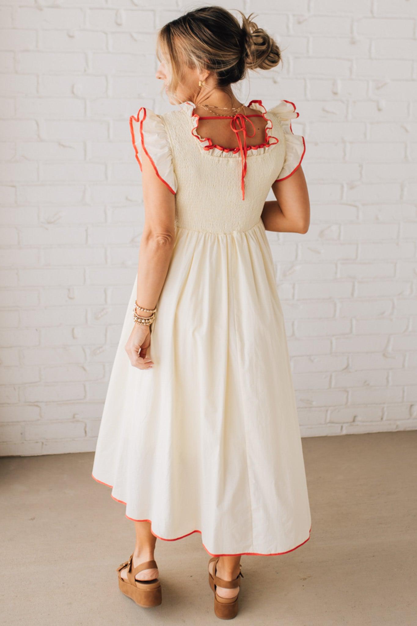 BLONDE MODEL WEARING A CREAM DRESS WITH RED CONTRASTED BINDING DETAIL AND SMOCKED BODICE MIDI DRESS WITH RUFFLE ACCENTS AND TIE BACK DETAIL.