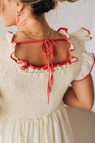 BLONDE MODEL WEARING A CREAM DRESS WITH RED CONTRASTED BINDING DETAIL AND SMOCKED BODICE MIDI DRESS WITH RUFFLE ACCENTS AND TIE BACK DETAIL.