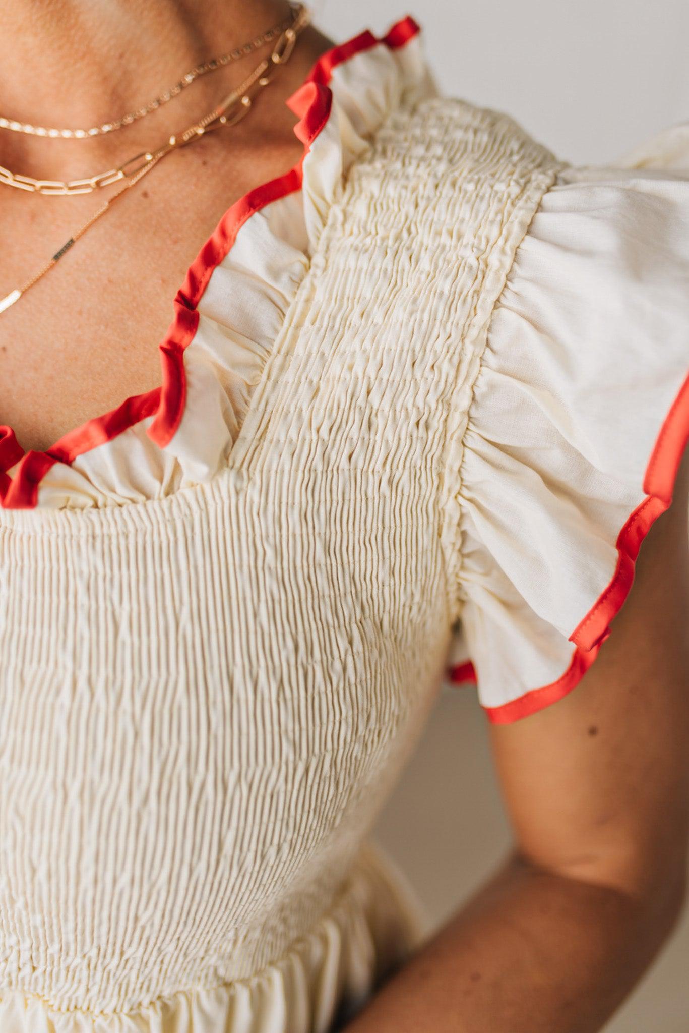 BLONDE MODEL WEARING A CREAM DRESS WITH RED CONTRASTED BINDING DETAIL AND SMOCKED BODICE MIDI DRESS WITH RUFFLE ACCENTS AND TIE BACK DETAIL.