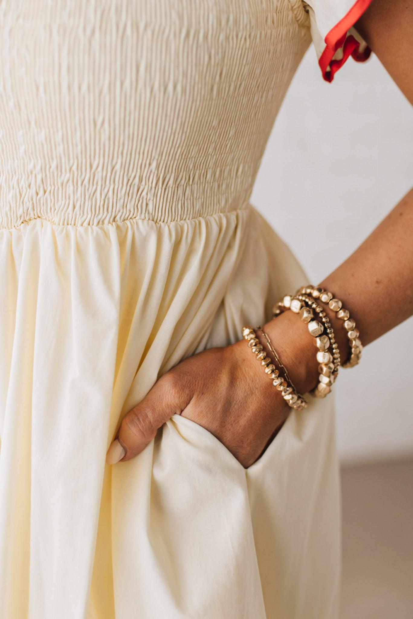 BLONDE MODEL WEARING A CREAM DRESS WITH RED CONTRASTED BINDING DETAIL AND SMOCKED BODICE MIDI DRESS WITH RUFFLE ACCENTS AND TIE BACK DETAIL.