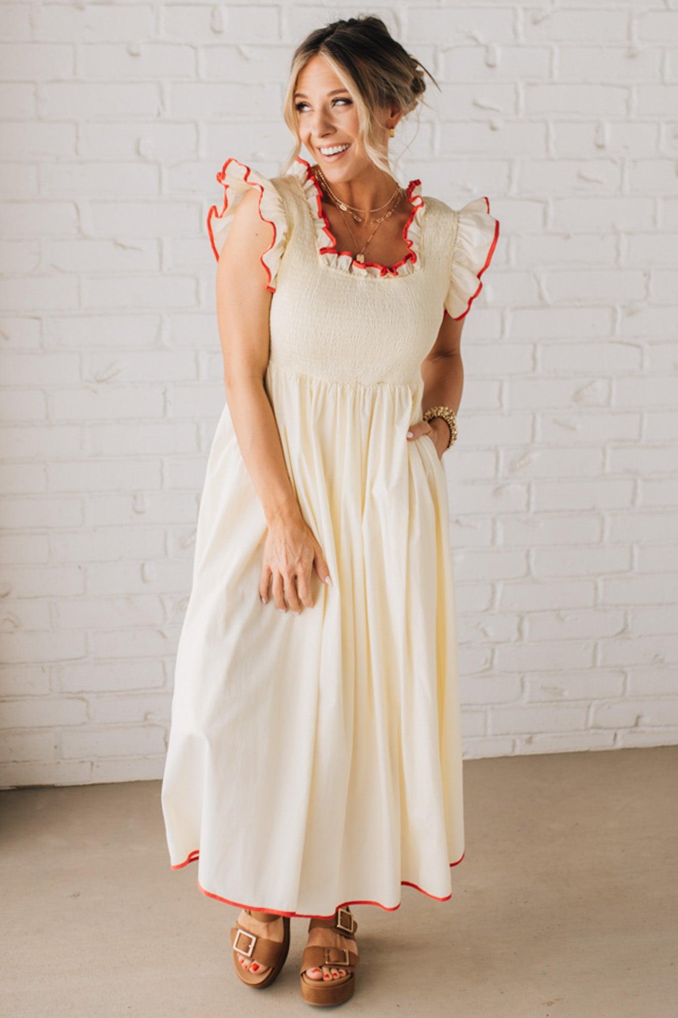 BLONDE MODEL WEARING A CREAM DRESS WITH RED CONTRASTED BINDING DETAIL AND SMOCKED BODICE MIDI DRESS WITH RUFFLE ACCENTS AND TIE BACK DETAIL.