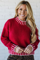 Woman wearing a red sweater with white trim against a white brick wall.