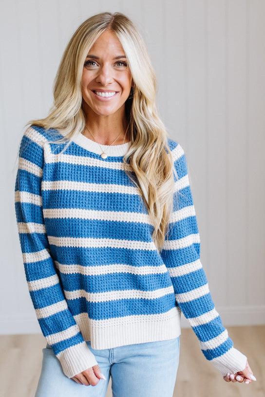 A smiling blonde woman wearing a blue and white horizontally striped knit sweater with light blue jeans, standing against a light neutral paneled wall.