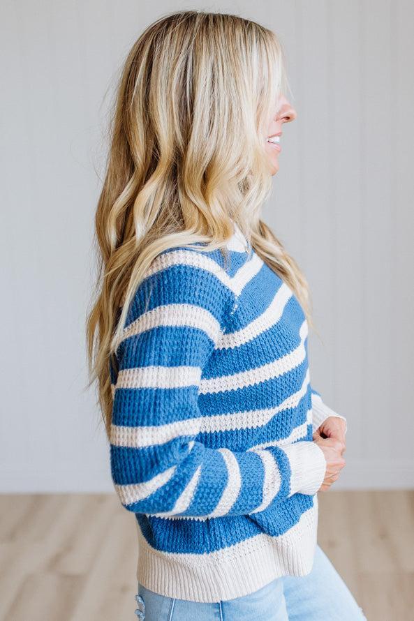 A woman in side profile wearing a blue and white striped knit sweater, paired with light blue jeans, standing in a brightly lit room with a light gray wall and wood floor.