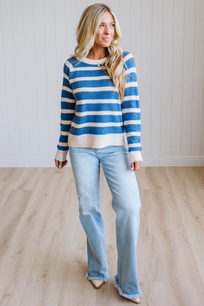 A woman modeling a blue and cream striped knit sweater with light-wash flare jeans, standing in a bright room with light wood floors and white paneled walls.