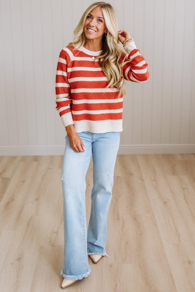 A woman modeling a red and white horizontally striped knit sweater with light-wash flare jeans and beige pointed-toe flats, standing on a light wood floor against a white paneled wall.