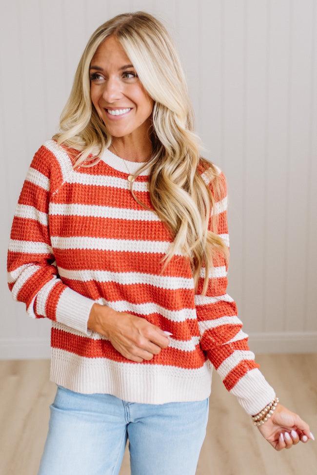 A smiling blonde woman wearing a waffle-knit crewneck sweater with horizontal rust orange and off-white stripes, paired with light blue jeans, standing against a light wood-paneled wall.