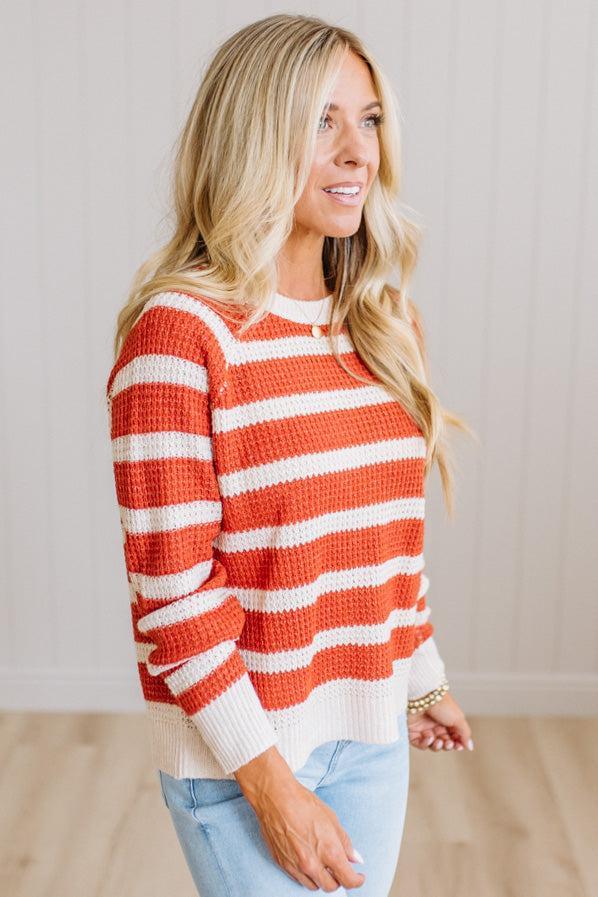 A woman wearing a waffle-knit crewneck sweater with horizontal orange and white stripes, paired with light blue jeans, standing against a light beige paneled wall.