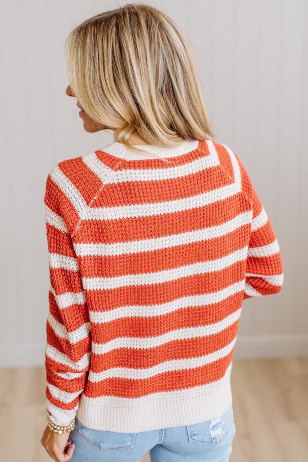 A woman viewed from behind wearing a waffle-knit sweater with horizontal orange and white stripes, paired with light blue jeans.