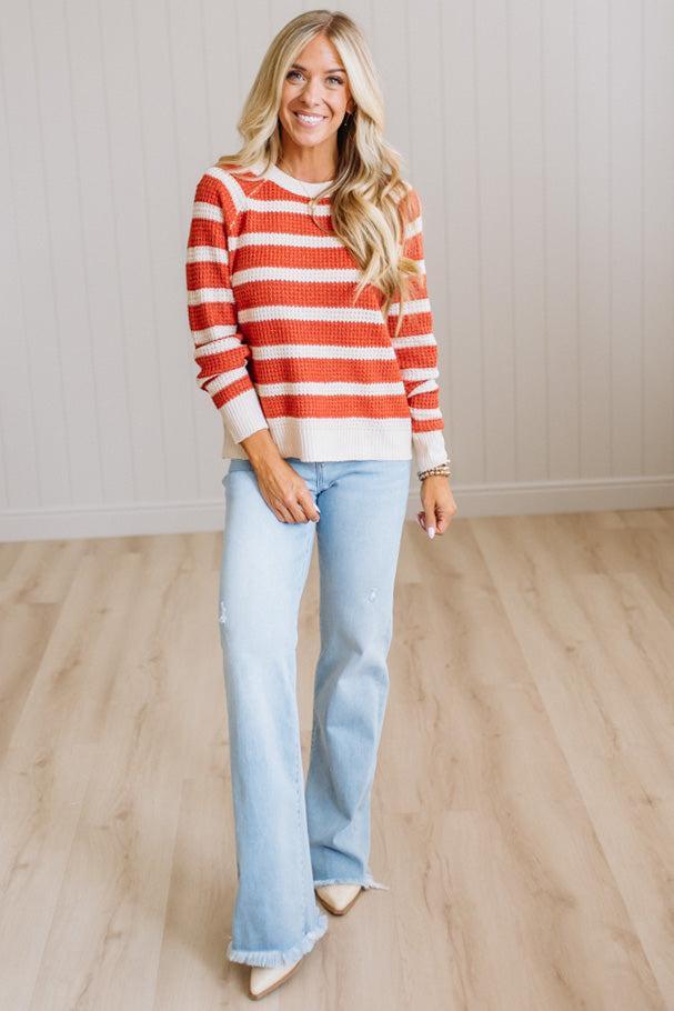 A woman stands in a bright room wearing a rust-orange and white striped knit sweater, light-wash blue flare jeans, and beige pointed-toe flats.