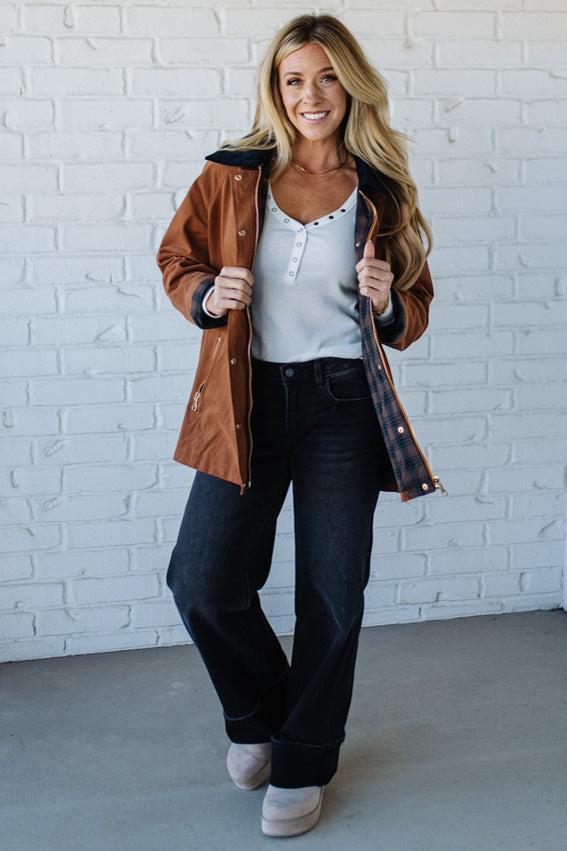 Woman wearing a brown jacket over a white shirt and black pants against a white brick wall.