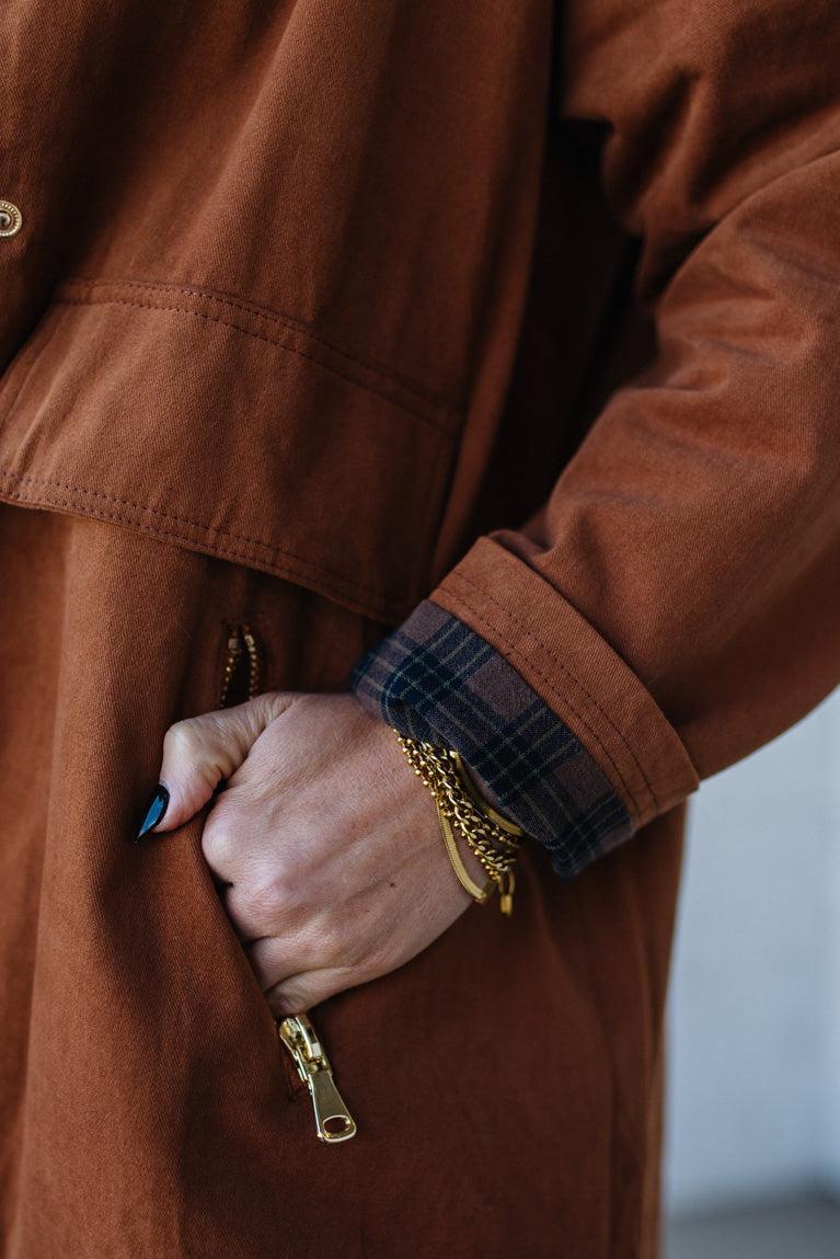 Close-up of a person wearing a brown coat with a plaid cuff and gold chain, holding a pen.