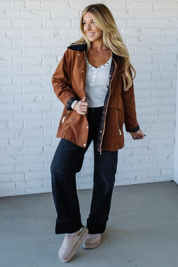 Woman wearing a brown jacket, white shirt, and dark jeans against a white brick wall.