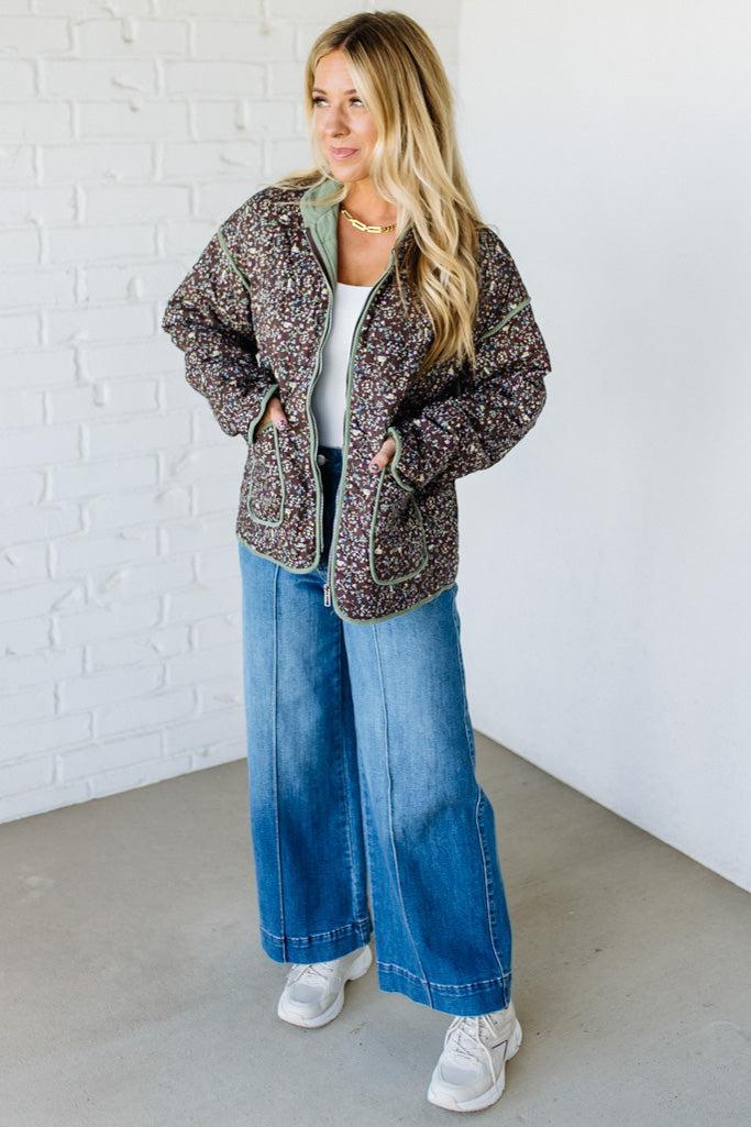 Woman wearing a patterned jacket, white shirt, blue jeans, and white sneakers against a white brick wall.