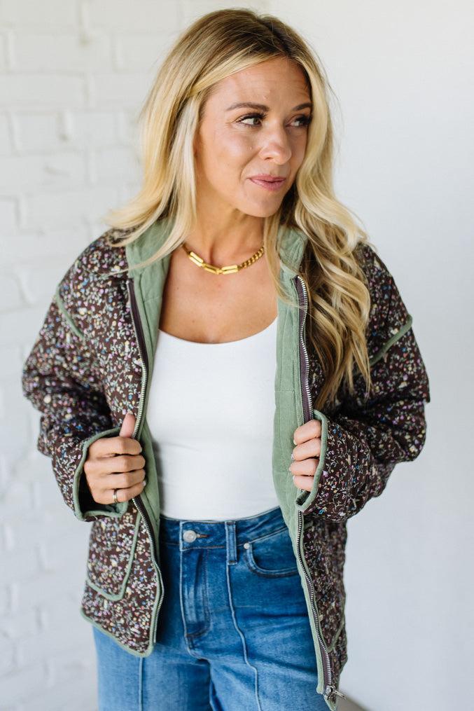 Woman wearing a floral jacket over a white top and blue jeans against a white brick wall.