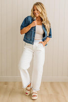 Woman wearing a denim jacket, white top, and white pants against a beige wall.