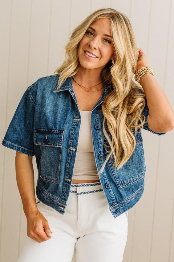 Woman wearing a denim jacket over a white top and pants, standing against a light wooden panel background.