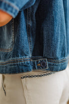 Close-up of a blue denim jacket with a button, worn over beige pants.
