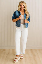 Woman wearing a denim jacket, white pants, and sandals indoors against a light wooden paneled wall.