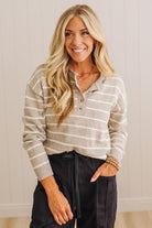 A smiling blonde woman wearing a beige and white horizontally striped long-sleeve henley top with dark charcoal pants, standing against a light beige paneled wall.