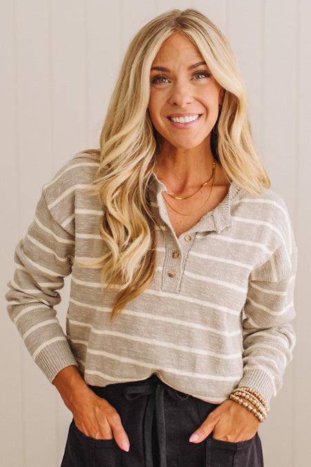 A smiling blonde woman wearing a beige and white horizontally striped long-sleeve knit polo shirt, paired with black pants and gold necklaces, standing against a light beige paneled wall.