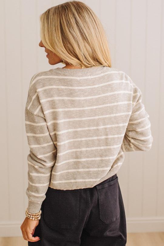A woman shown from the back wearing a beige and white horizontally striped knit sweater and dark pants, standing against a light paneled wall.