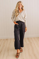A woman modeling a beige and white striped long-sleeve polo shirt paired with dark charcoal cropped pants and brown leather slide sandals, standing in a room with light wood floors and white paneled walls.