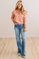 A woman modeling a long-sleeve, collared knit top with horizontal peach and white stripes, paired with blue jeans and beige pointed-toe heels, standing in a room with light wood floors and white paneled walls.