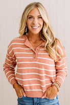 A smiling blonde woman wearing a long-sleeve knit polo sweater with horizontal peach and white stripes, paired with blue jeans and layered gold bracelets, against a light neutral background.