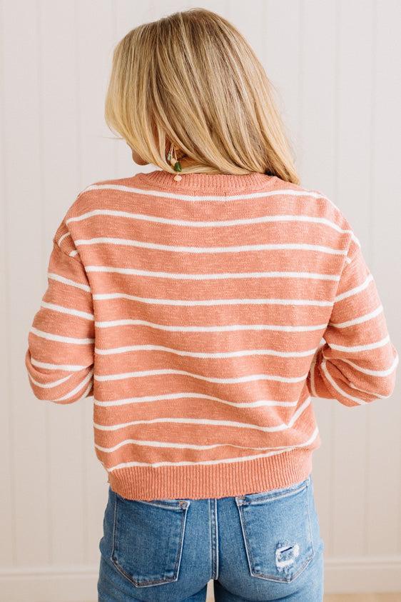 A woman viewed from behind wearing a coral pink and white horizontally striped knit sweater with blue jeans, set against a light beige paneled wall.